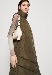 Gilet matelassé vert olive avec col haut, fermeture zip et poches latérales. Le modèle tient un petit sac à main blanc avec une bandoulière décorative.