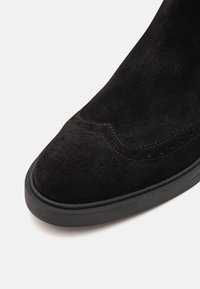 Svart mockaskor med rund tå, med en slät yta och dekorativ söm längs sömmen. Gummisula med texturerad kant.