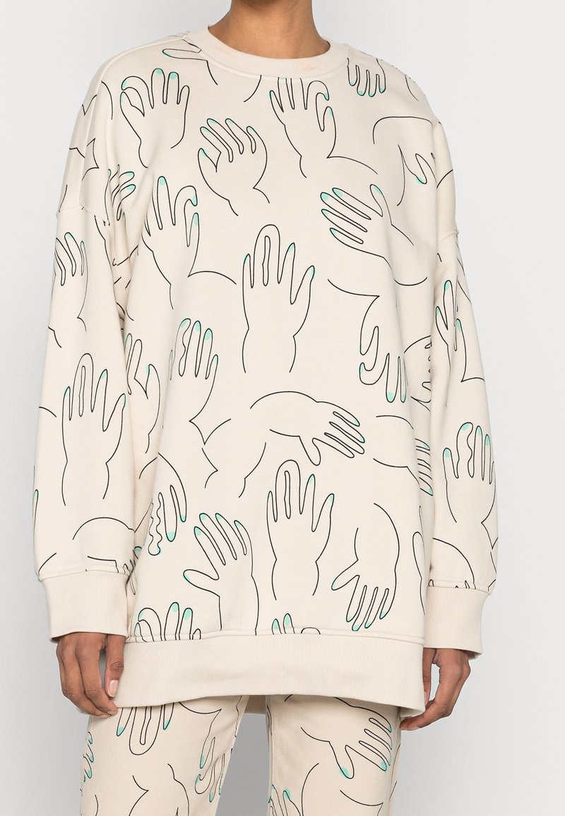 Crèmekleurige sweatshirt met zwarte lijn-tekeningen van handen en teal accenten. Heeft een losse pasvorm, lange mouwen en ribgebreide boorden aan de onderkant.