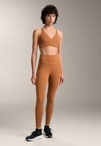 Rjav, raztegljiv športni crop top in hlače z visokim pasom, ki imata gladko teksturo z minimalnimi šivi; v kombinaciji s črnimi športnimi čevlji.