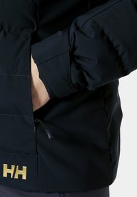 Navy Pufferjacke mit glatter Textur, ausgestattet mit gesteppten Details, einer Reißverschlusstasche und einem goldenen Logoakzent an der Unterseite.
