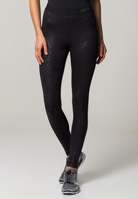 Leggings neri con un sottile motivo in pelle di serpente, realizzati in materiale elasticizzato. Presentano una vita alta e un design aderente, abbinati a sneaker grigie.