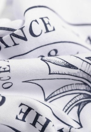 Weißer Stoff mit einem schwarzen Aufdruck, der komplexe Linien und Text zeigt, wodurch ein strukturiertes Erscheinungsbild mit sichtbaren Falten und knitterungen entsteht.