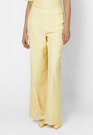 Woman wearing high-waisted, wide-leg, pale yellow trousers paired with matching pointed-toe shoes, standing against a white background.