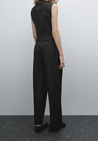 Vrouw in een mouwloze zwarte top, zwarte wijde broek en zwarte fuzzy pantoffels, staand tegen een effen lichtgrijze muur.