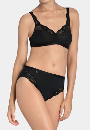 Mujer con un sujetador negro con encaje y una braguita de corte alto a juego, contra un fondo claro y liso.