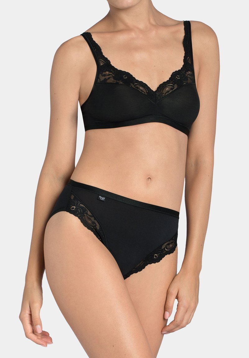 Kobieta w czarnym biustonoszu zdobionym koronką i dopasowanych majtkach o wysokim kroju na jednolitym jasnym tle.