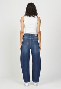 Weite, dunkelblaue Denim-Jeans mit lockerer Passform, ausgestattet mit zwei Gesäßtaschen und einem beigefarbenen Etikett am Bund. Das Model trägt ein weißes Trägertop.