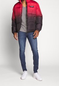 Chaqueta de plumas roja y negra con bolsillos con cremallera, puños acanalados y logotipo. Llevada sobre una camiseta gris de cuello en V y combinada con jeans azules y zapatillas blancas.