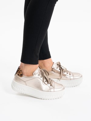 Persoon draagt metalen beige plateau sneakers met luipaardprint accenten op de hiel en beige veters, gecombineerd met zwarte nauwsluitende broek.
