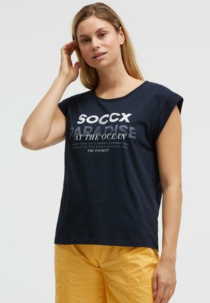 Frau mit langen blonden Haaren, die ein ärmelloses navyblaues Shirt mit Text und senfgelben Hosen trägt, steht vor einem weißen Hintergrund.