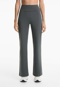 Model trägt graue, hoch geschnittene Yogahosen mit einer figurbetonten Silhouette und weißen Sportschuhen, vor einem schlichten weißen Hintergrund.