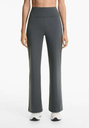 Modèle portant un pantalon de yoga haut à taille haute gris avec une silhouette ajustée et des chaussures de sport blanches, sur fond blanc uni.