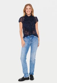 Blusa trasparente blu navy con dettagli a volant e maniche corte, abbinata a jeans azzurri e mocassini neri, in piedi di fronte a uno sfondo bianco.
