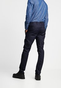 Dark blue denim pants with a slim fit, featuring back pockets and a smooth texture. Paired with a light blue button-up shirt.