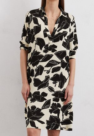 Vrouw draagt een roomkleurige jurk met grote abstracte zwarte bloemenprint, opgerolde mouwen en een V-hals.