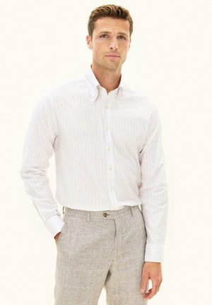 Mężczyzna w białej koszuli na guziki z cienkimi paskami i jasnoszarych spodniach w kratę, stojący z jedną ręką w kieszeni na białym tle.