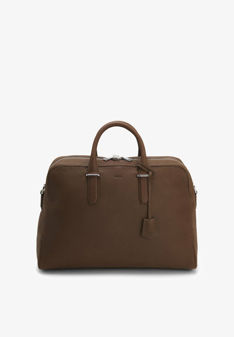 Sac de voyage en cuir marron avec doubles poignées, fermeture éclair argentée et petite étiquette en cuir attachée sur fond blanc.