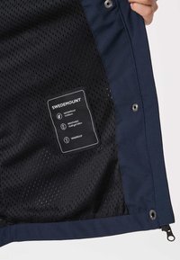 Roka, ki drži odprt temnomodri suknjič, ki razkriva črto mrežaste podloge z etiketo Swedemount, na kateri piše, da je vodoodporen, zračen in vetrovno odporen.