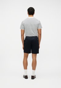 Άνδρας που στέκεται με την πλάτη στην κάμερα, φοράει λευκό και μαύρο ριγέ μπλουζάκι, σκούρα μπλε σορτς, λευκές κάλτσες και σκούρα καφέ παπούτσια.