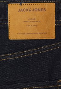 Mörkblå denimtyg med gul sömnad. En brun läderetikett med präglad text som detaljerar "JACK & JONES, JEANS INTELLIGENCE, SEDAN 1990."