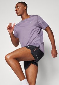 Lilla atletisk t-shirt med korte ærmer, parret med sorte shorts med mønstret tekstur, orange accenter og reflekterende detaljer.