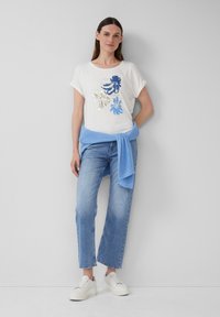 Witte T-shirt met blauwe en zilveren bloemenontwerpen, gecombineerd met een lichtblauwe gebreide trui om de taille en lichtdenim jeans. Witte sneakers.