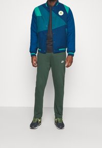 Man som bär en blå och teal varsityjacka, mörkgrå tröja, gröna Nike sweatpants och svarta sneakers med gröna snören, står mot en vit vägg.