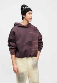 Sweatshirt roxa com design desgastado, apresentando rasgos e um bolso frontal. O modelo veste um gorro preto com letras brancas e calças creme.