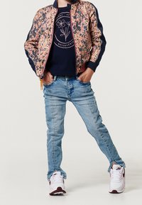 Bambino che indossa una giacca a fantasia rosa e blu navy, una camicia blu navy con grafica floreale, jeans azzurri e sneakers bianche Nike, in piedi con le mani nelle tasche.