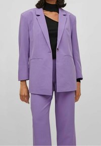 Costume deux pièces violet comprenant un blazer cintré à un bouton, avec des revers crantés, et un pantalon à jambes larges. Le tissu a une texture lisse.