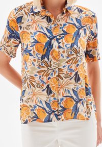 Collared short-sleeve blouse featuring a cream background with vibrant orange and blue floral and fruit prints. Made of lightweight fabric.