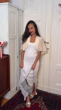 Femme aux longs cheveux foncés portant une robe blanche à motifs, un court châle beige, des sandales argentées et tenant un sac à main argenté, debout à l'intérieur.