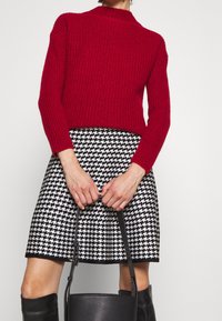 Punainen ribbineule neule yhdistetty mustavalkoiseen tikattuun hameeseen. Kädessä pidettävä musta laukku, polven korkuiset mustat saappaat, teksturoitu kangas.