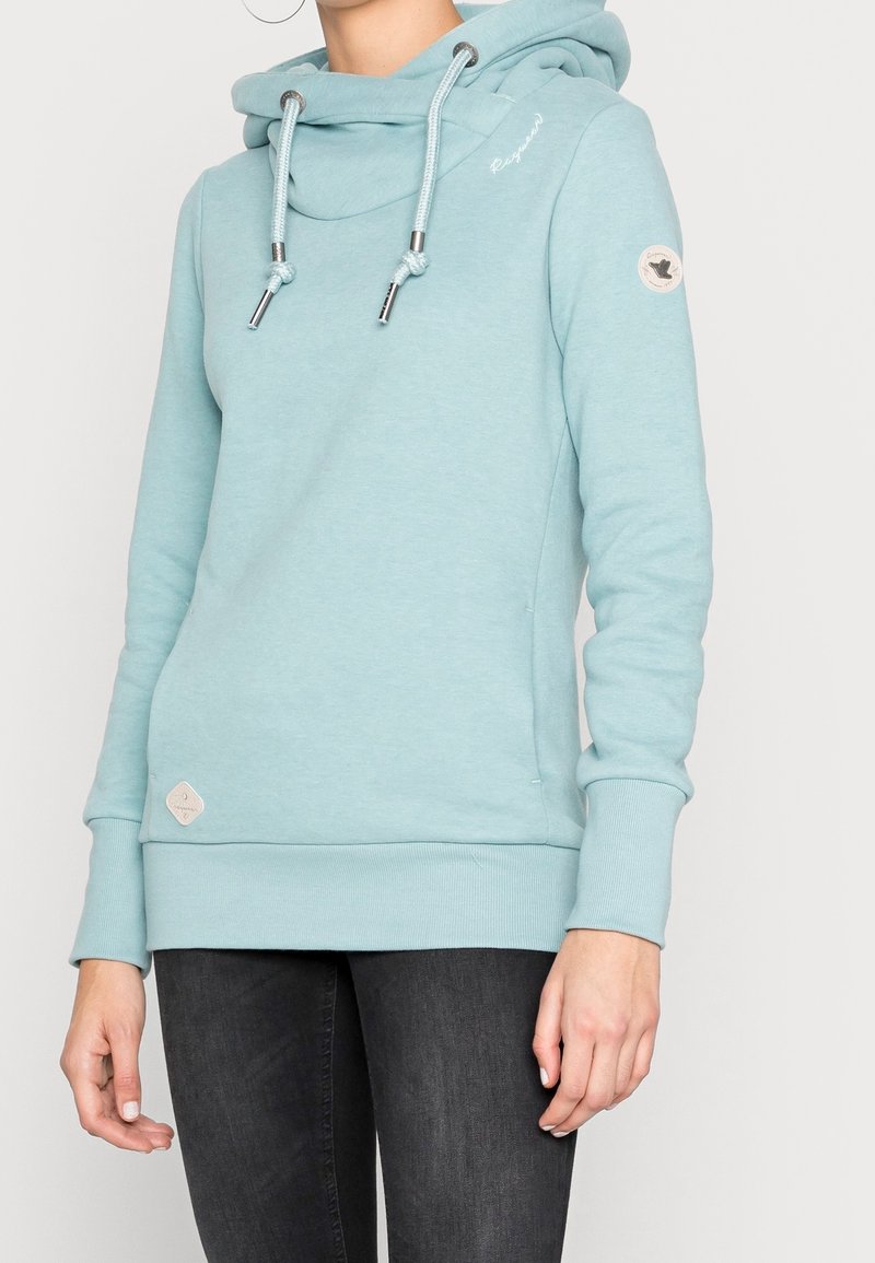 Ljusblå hoodie med cowl-krage, elastiska mudd och fåll. Har metalliska dragsko och ett märke på vänster ärm. Slät textur.