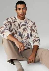 Cream knitted sweater featuring a multicolored abstract pattern in brown and blue, paired with beige trousers and light gray textured socks.