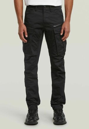 Zwarte cargobroek met meerdere zakken en ritssluiting, gedragen met zwarte sneakers en een wit overhemd, getoond van taille tot voeten.