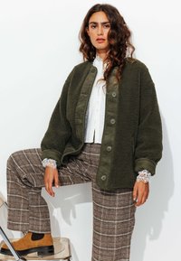 Veste polaire vert olive à boutons sur le devant avec détails en velours côtelé, associée à un pantalon ample à carreaux, sur un fond blanc.