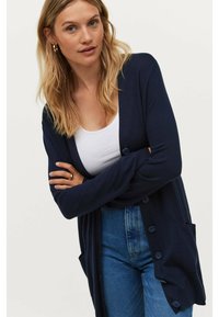 Marineblauer, langer Cardigan aus weichem Stoff, mit V-Ausschnitt, vorderen Knöpfen und zwei Seitentaschen. Kombiniert mit einem weißen Oberteil und Jeans.