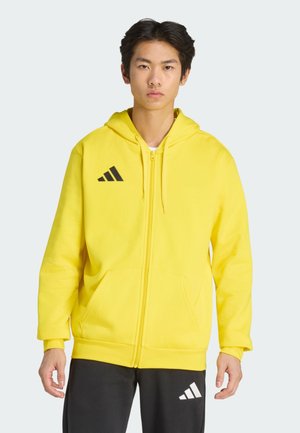 Junger Mann trägt einen knallgelben Hoodie mit Reißverschluss und schwarzem Adidas-Logo sowie schwarze Hose mit weißem Adidas-Logo, stehend vor einfachem Hintergrund.