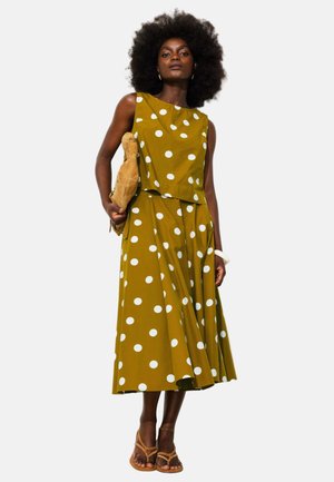 Femme avec une coiffure afro portant une robe sans manches moutarde à pois blancs, tenant une pochette beige et portant des sandales.