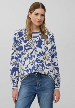 Blouse florale en bleu et crème avec un décolleté en V, des manches longues et une fermeture à boutons, superposée sur un haut rayé bleu marine et blanc. Portée avec un jean.
