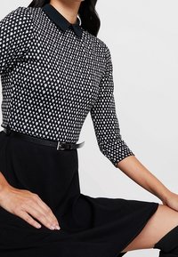 Robe noire avec un corsage ajusté, des manches texturées à motif noir et blanc, un col noir et une fine ceinture noire à la taille.