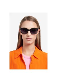 Ochelari negri cu rame mari și rectangulare și lentile întunecate, având accente metalice. Modelul poartă o cămașă portocalie peste un top roz.