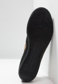 Schwarze Sneaker-Sohle mit geometrischem Profilmuster, die an einer weißen Eckfläche lehnt, mit feinen Text- und Logodetails.