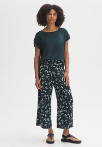 Donkergroene blouse met korte mouwen gecombineerd met zwarte wijde broek met een mintgroen abstract patroon. Zwarte platte sandaaltjes.