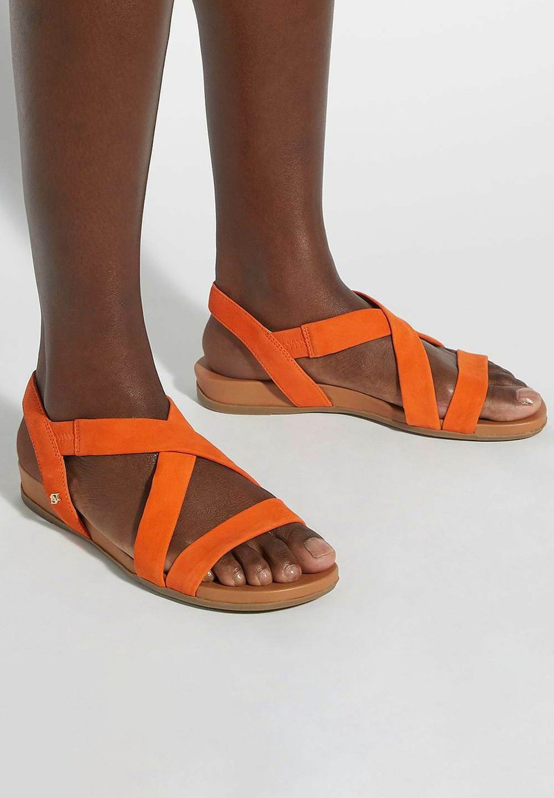 Orange suede sandals with crisscross straps, open toes, and a low rubber sole. Features a small decorative logo on the side.