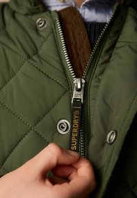 Zaļš quiltēts jaku ar sudraba rāvējslēdzēju un spiedpogu. Melna un dzeltena rāvējslēdzēja vilkšanas teksts, kas lasāms "SUPERDRY". Redzami iekšējie slāņi.