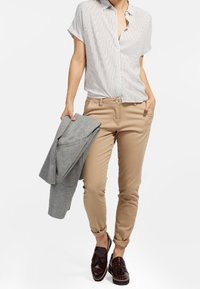 Tanové chinos s ohrnutými manžetami, spárované s krátkým rukávem, bílou a šedou pruhovanou košilí s knoflíky. Šedý blejzr položený na jedné paži. Hnědé boty.