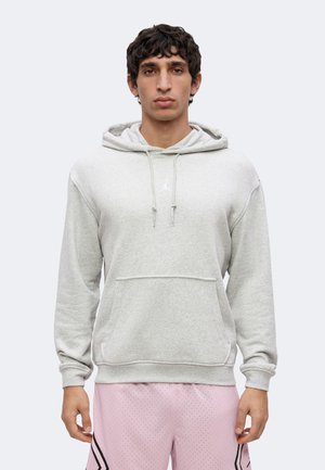 Jeune homme aux cheveux bouclés et foncés portant un sweat à capuche gris clair et un short de basket rose avec des rayures noires.